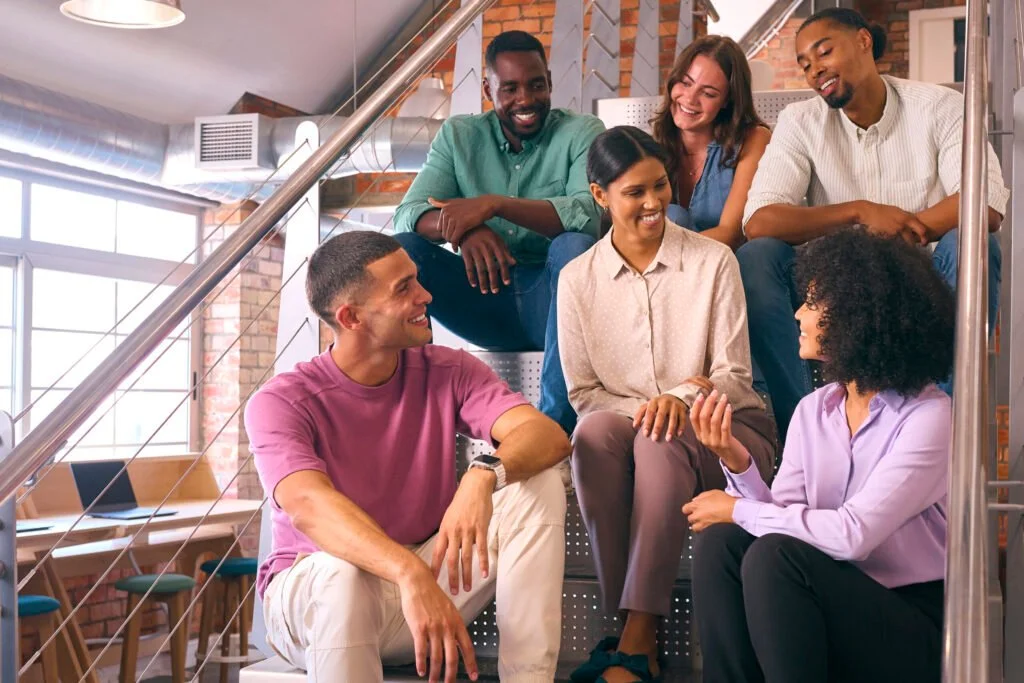 Inspiring Stories from Local Heroes smiling multi cultural business team sitting on stairs in modern open plan office