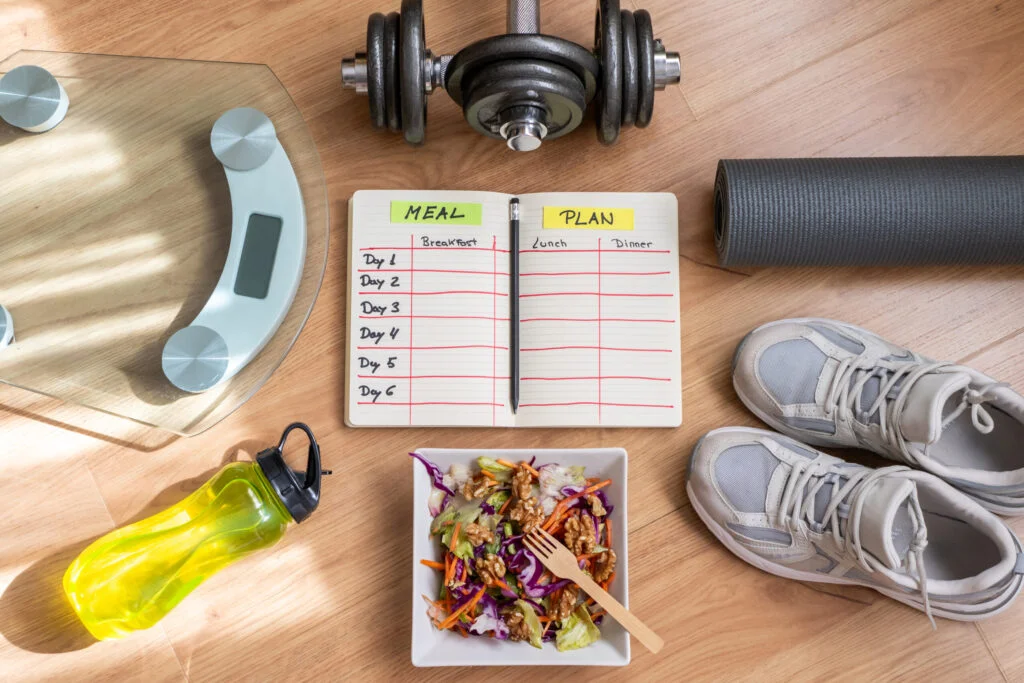 overhead view of a wooden floor featuring dumbbells. a yoga mat. sneakers. a scale. a water bottle. a meal plan notebook. and a healthy salad